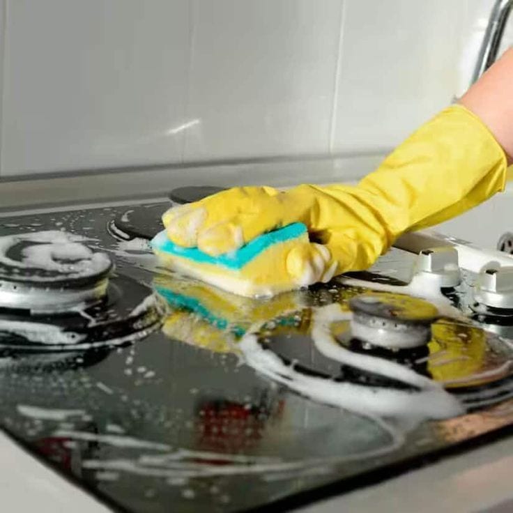 A gloved hand scrubbing a soapy glass stovetop